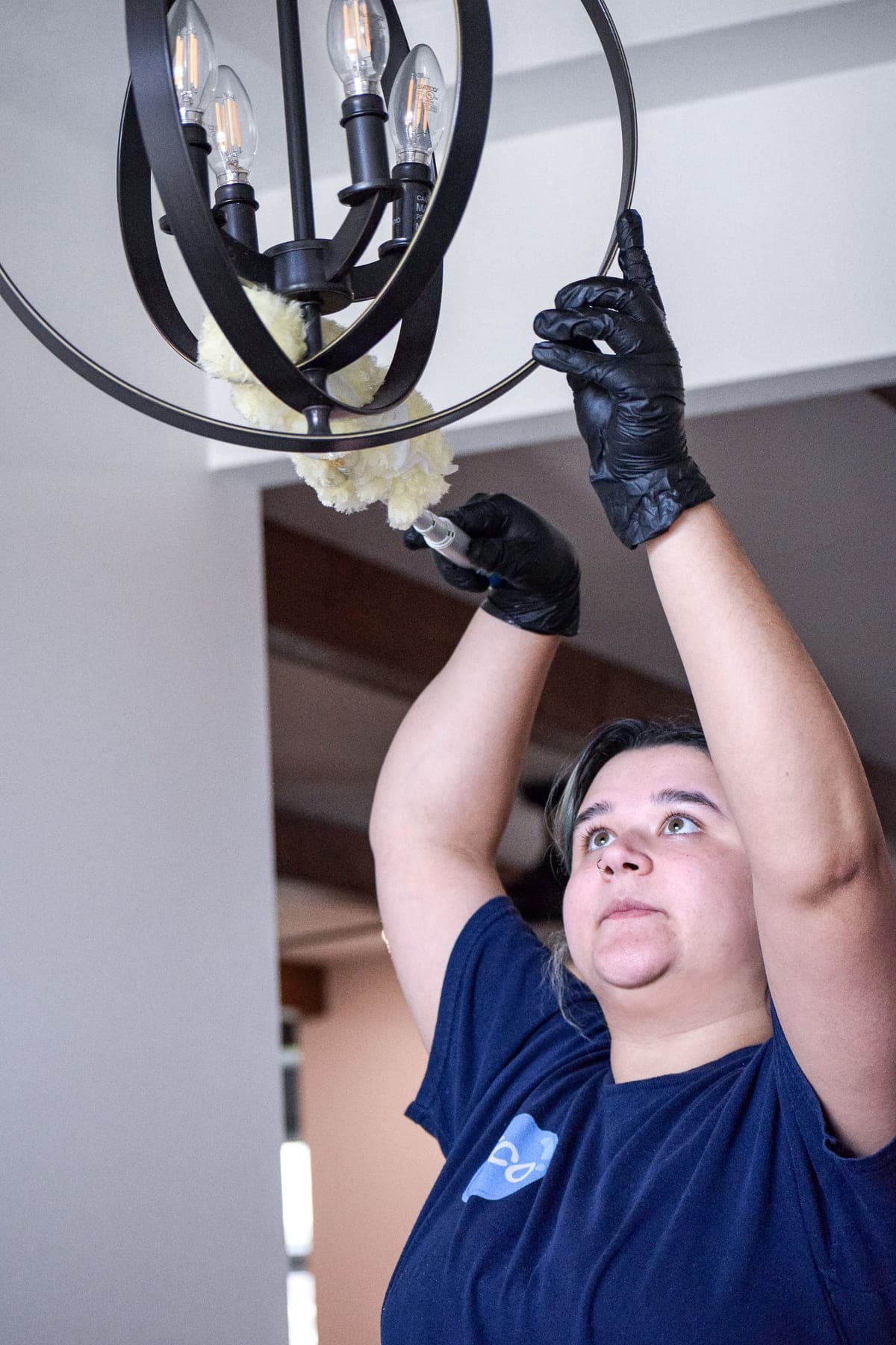 An ECO cleaner reaching overhead to dust the arms of a black orb chandelier with a duster.