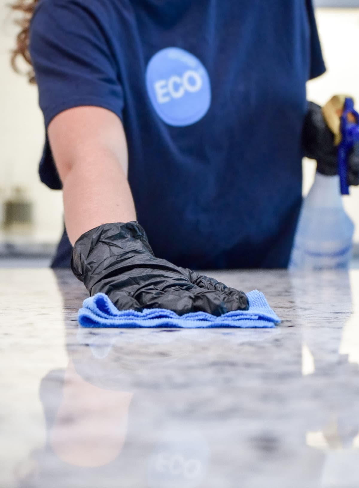 A gloved ECO cleaner wiping down a polished granite countertop with a blue microfiber cloth, spray bottle in the other hand.