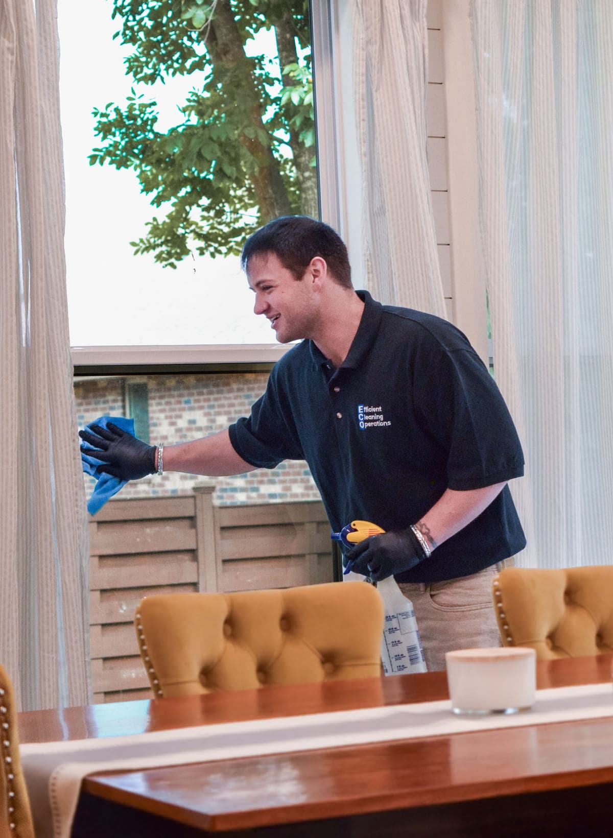 An ECO cleaner in a company polo wiping a window beside a dining room table.