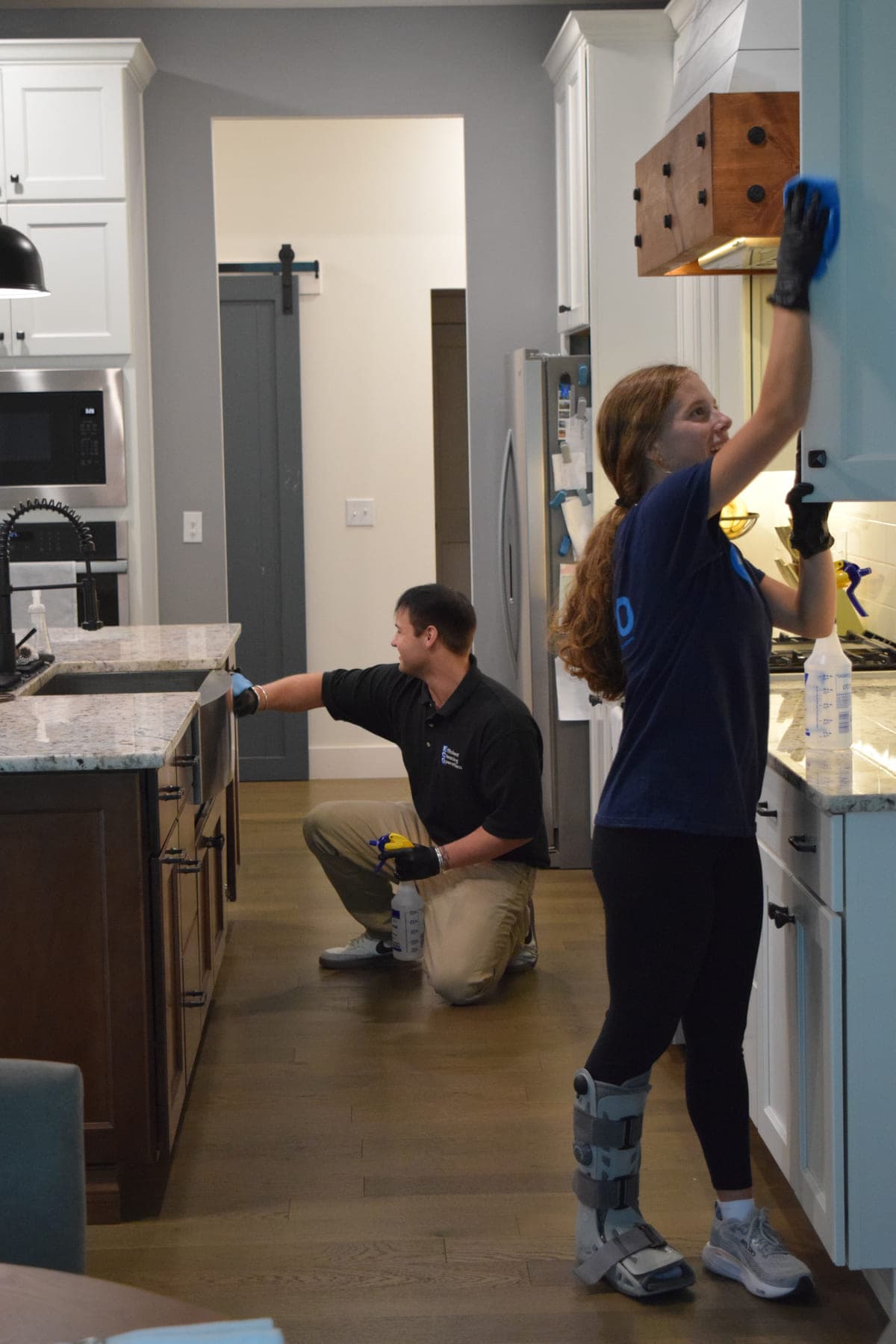 Two ECO cleaners in a kitchen — one reaching up to wipe the wood range hood, the other kneeling at the island to clean a cabinet front.