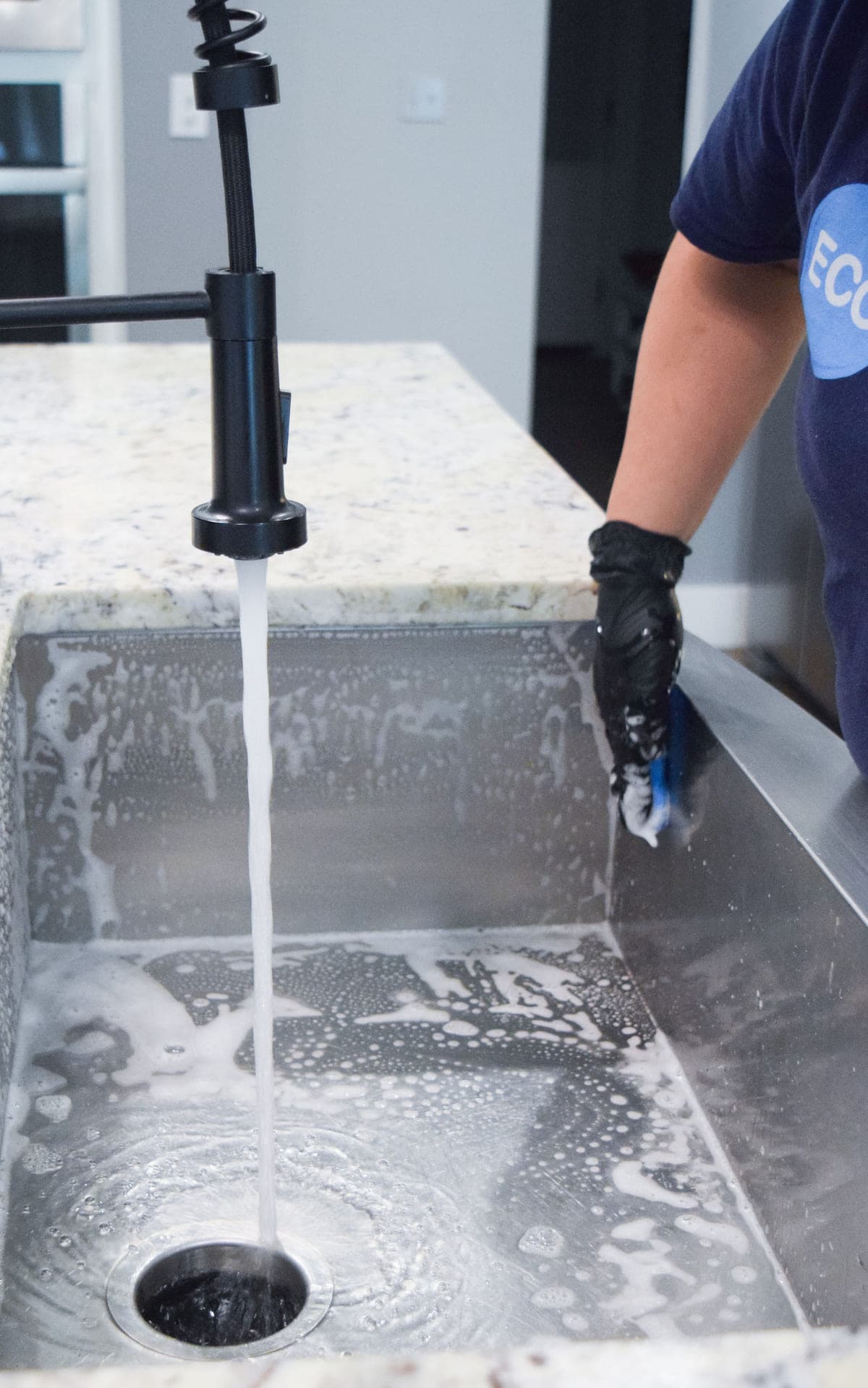 A gloved ECO cleaner scrubbing a stainless steel kitchen sink, water running and suds across the basin.