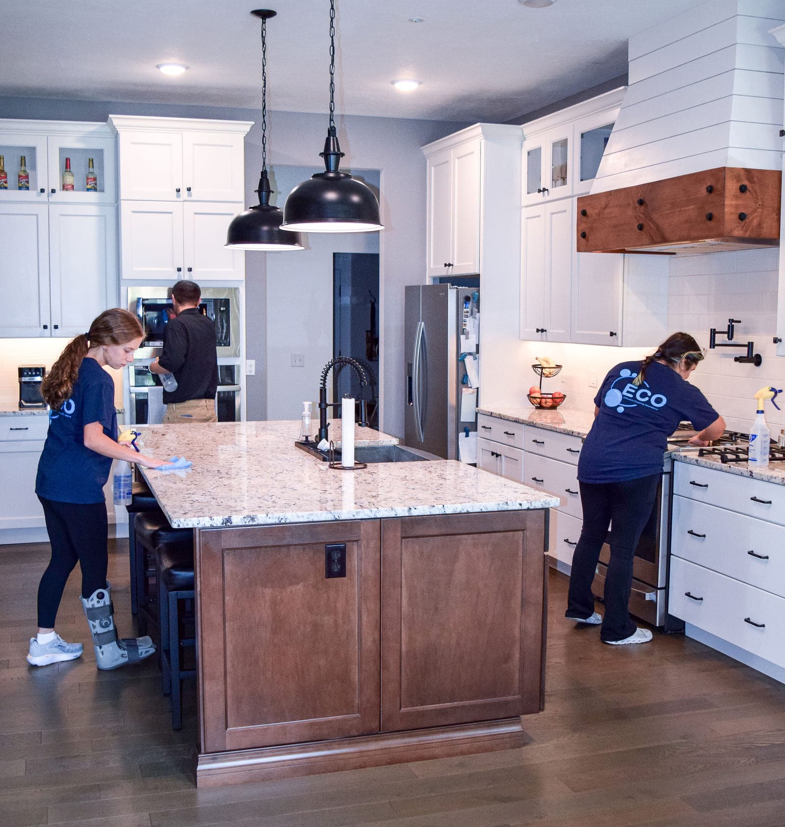Three ECO cleaners at work in a bright kitchen — one wiping down the granite island, one at the wall oven, one cleaning the counter beside the range.